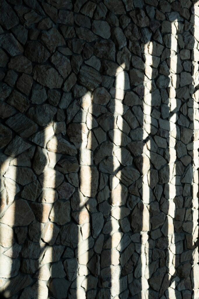 Photo by HYEWON HWANG Shadows of vertical bars cast on a stone wall.